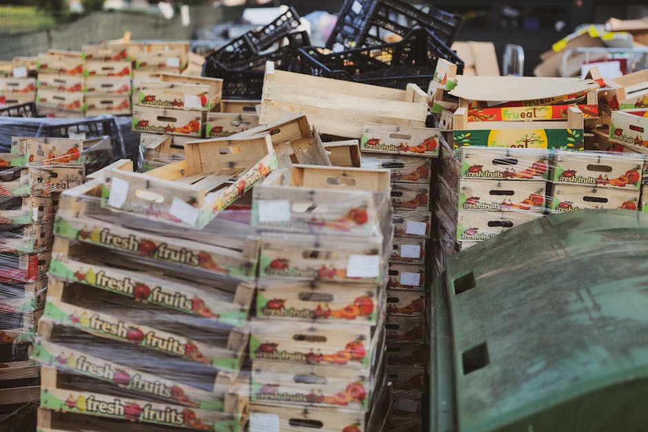 A large outdoor scene featuring multiple stacks of cardboard fruit and vegetable boxes, primarily labeled 'fresh fruits' with colorful images of tomatoes and other produce. The boxes are arranged in uneven stacks, some leaning or partially collapsed, and are placed on a paved surface, likely a street or market area. Behind the boxes, there are black plastic crates, some piled atop each other, and a few wooden pallets. To the right, a weathered green container or bin with visible scratches and dirt marks is partially visible, suggesting it is used for waste collection or storage. The overall environment appears to be a busy outdoor market or warehouse loading area where goods are being cleared or prepared for disposal, fitting with the context of rubbish removal or waste handling, as managed by Waste Clearance Ruislip or similar services.