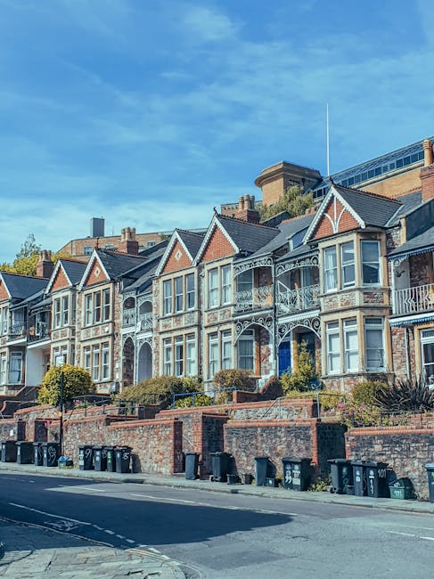 A row of Victorian-style terraced houses with ornate decorative ironwork and bay windows, situated on a gentle slope next to a paved street. The houses feature brick and painted finishes in neutral tones, with some elements in white and light grey, and pitched roofs covered with dark tiles. Small front gardens with neatly trimmed bushes and low brick walls separate the properties from the pavement. Several black wheelie bins are lined up along the curb, indicating waste collection points typical of private waste management. In the background, modern office or apartment buildings are visible behind the historic houses, set against a clear blue sky. The scene represents urban residential architecture with a subtle connection to waste and rubbish removal services in a typical British neighborhood.