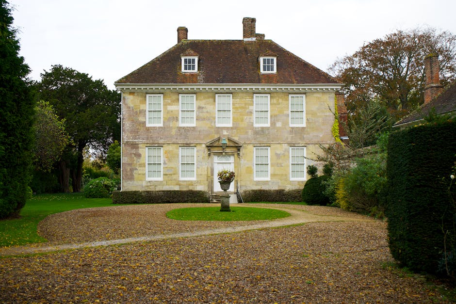 A large, historic stone house with a symmetrical façade, featuring three rows of white sash windows, a centrally located main door with decorative stonework, and a pitched roof covered in reddish-brown tiles. The front garden has a gravel driveway that curves around a small circular lawn with a stone birdbath or urn in the center, surrounded by neatly trimmed hedges and lush green lawns. Mature trees with sparse autumn foliage are visible to the sides and behind the house, creating a natural backdrop. The environment appears well-maintained and quiet, with the scene suggesting a private residential property. The overall setting conveys a sense of traditional British architecture, suitable for private waste collection or on-site clearance services by Waste Clearance Ruislip, in the context of rubbish removal or estate clearance.