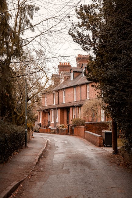 A row of Victorian-style terraced houses with ornate decorative ironwork and bay windows, situated on a gentle slope next to a paved street. The houses feature brick and painted finishes in neutral tones, with some elements in white and light grey, and pitched roofs covered with dark tiles. Small front gardens with neatly trimmed bushes and low brick walls separate the properties from the pavement. Several black wheelie bins are lined up along the curb, indicating waste collection points typical of private waste management. In the background, modern office or apartment buildings are visible behind the historic houses, set against a clear blue sky. The scene represents urban residential architecture with a subtle connection to waste and rubbish removal services in a typical British neighborhood.